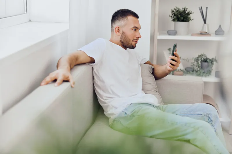 Thoughtful man checking phone while sitting on a sofa, reflecting on his digital detox journey and work-life balance, amidst a calming home environment.