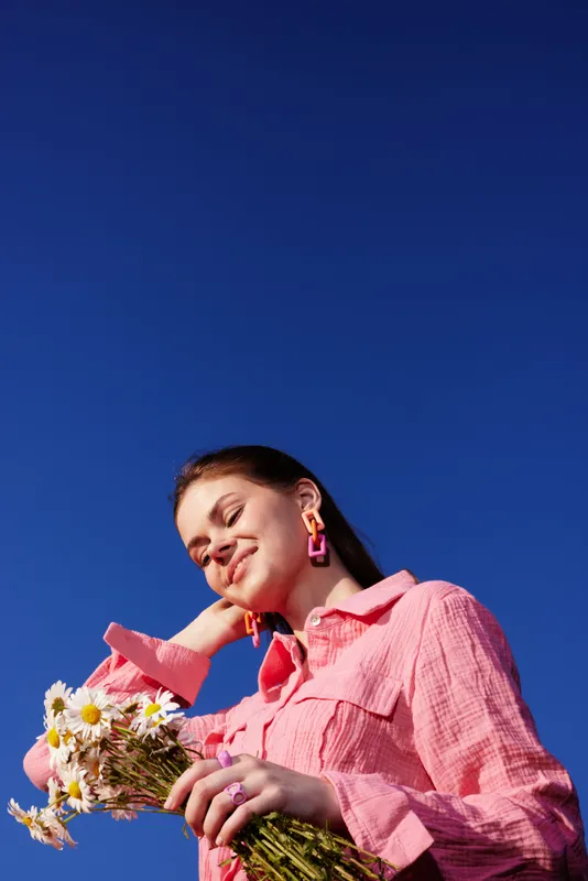 Young woman in pink shirt holding flowers against a blue sky, showcasing joy and freshness in a vibrant, seasonal outdoor setting