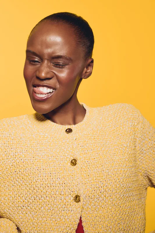 Young African woman with dark skin wearing a yellow knitted cardigan poses with eyes closed and tongue out. Studio portrait on yellow background, concept for joy, fashion, and self-expression.