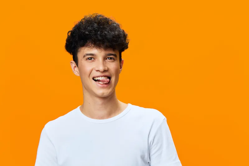 smiling young man in white t-shirt posing playfully with tongue out on vibrant orange background, cheerful expression, casual style, studio portrait
