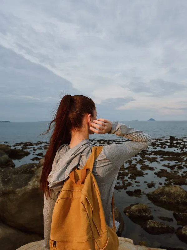 Young woman with a backpack enjoying a coastal view, wearing a gray hoodie, showcasing a sense of adventure and tranquility on a cloudy day