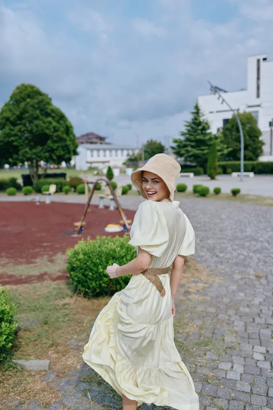 Woman in a light dress and straw hat walks outdoor in a landscaped garden, smiling, capturing a carefree summer moment as her dress swirls beside trimmed hedges