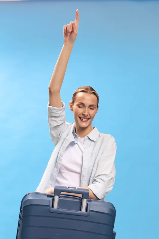 Happy woman with suitcase raises hand with finger up smiling on blue background. Concept for travel, vacation, success and positive emotions in lifestyle.