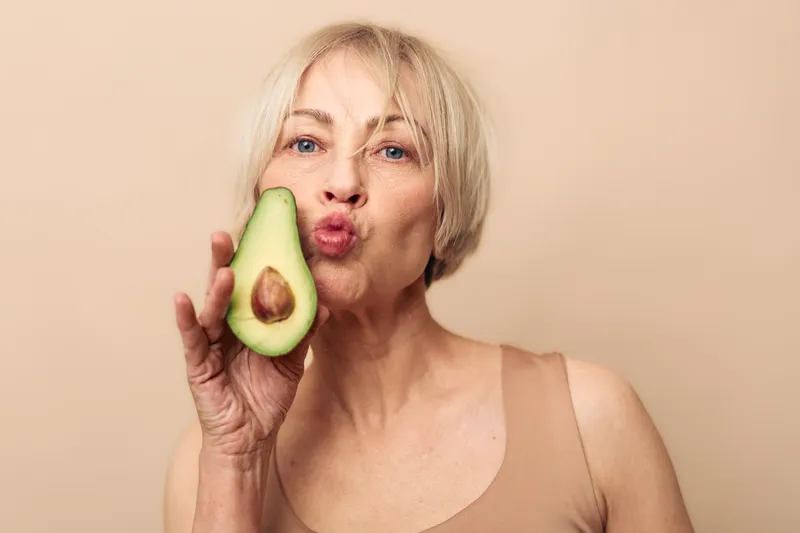 Mature woman with short blonde hair holds half an avocado near her face making a kiss gesture. Concept of healthy eating, natural beauty, skincare, and wellness on beige background.