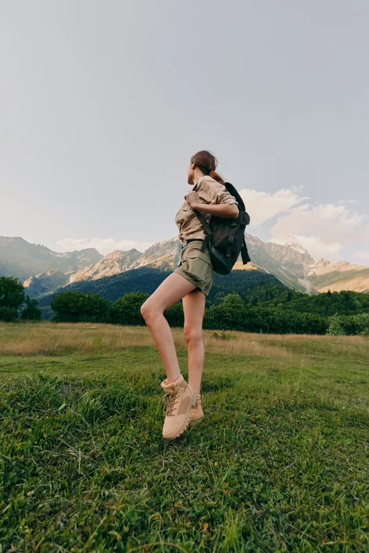 A woman jumps in a grassy field with a backpack against distant mountains, embodying outdoor adventure, lifestyle, nature, freedom, joy, energetic spirit in daylight and fresh air.