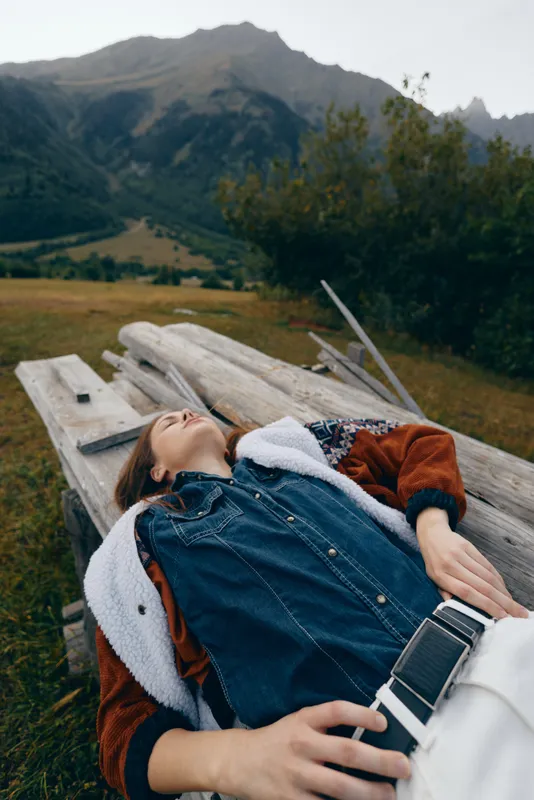 Woman mountains nature relax wooden meadow scene with young traveler lying on rustic bench in denim jacket, peaceful outdoor rest and scenic countryside landscape under cloudy sky