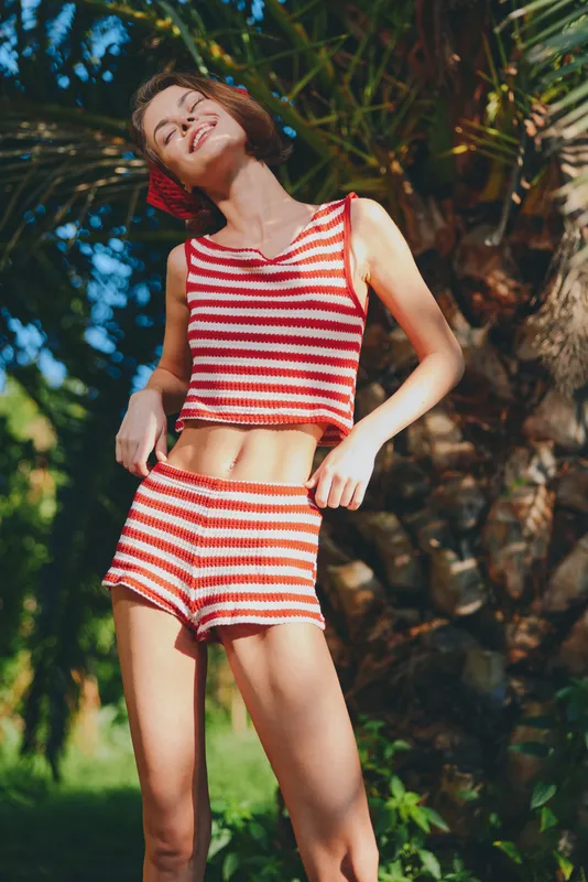 A cheerful woman wearing a red striped crop top and shorts poses outdoors in bright sunlight, her smile radiant as she enjoys a playful summer moment.