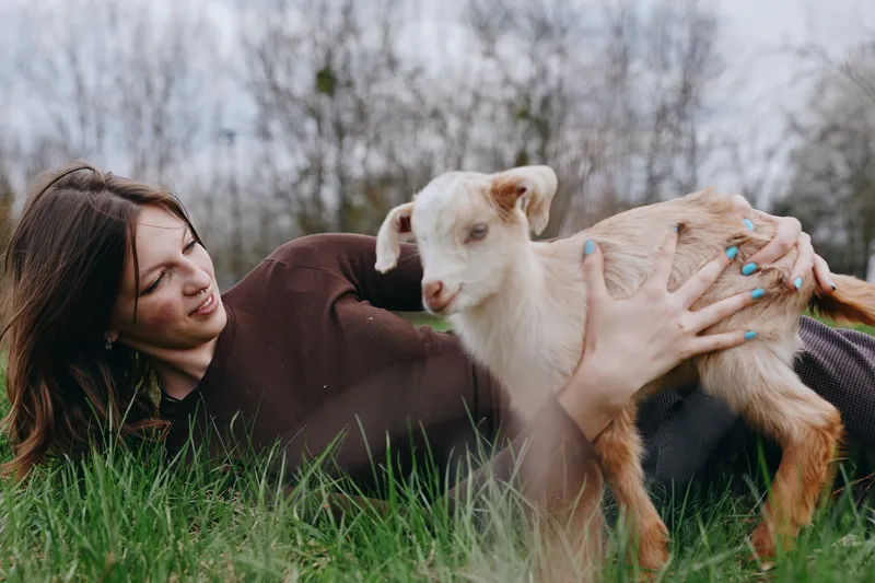 Young woman lying on grass gently holding a baby goat outdoors in a natural setting during a relaxed lifestyle moment.