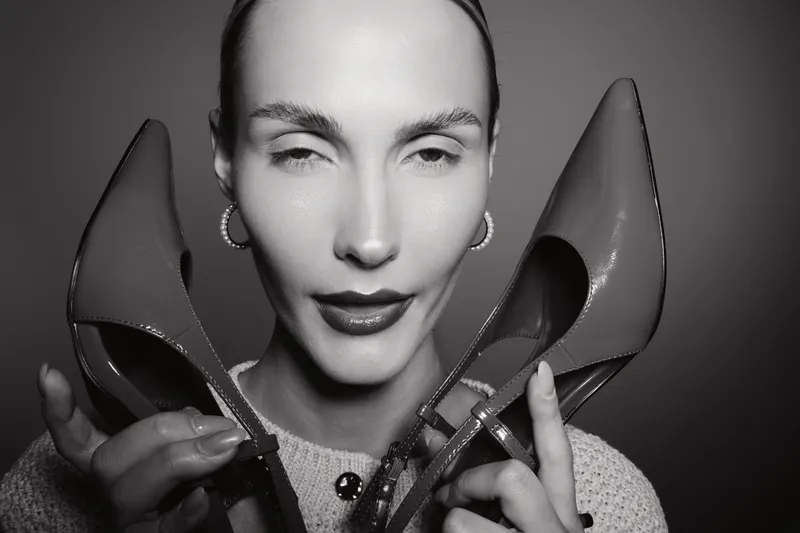 Close-up portrait of a fair-skinned Caucasian woman with slicked-back hair holding two high heel shoes near her face in black and white. Concept of fashion, style, and elegance.