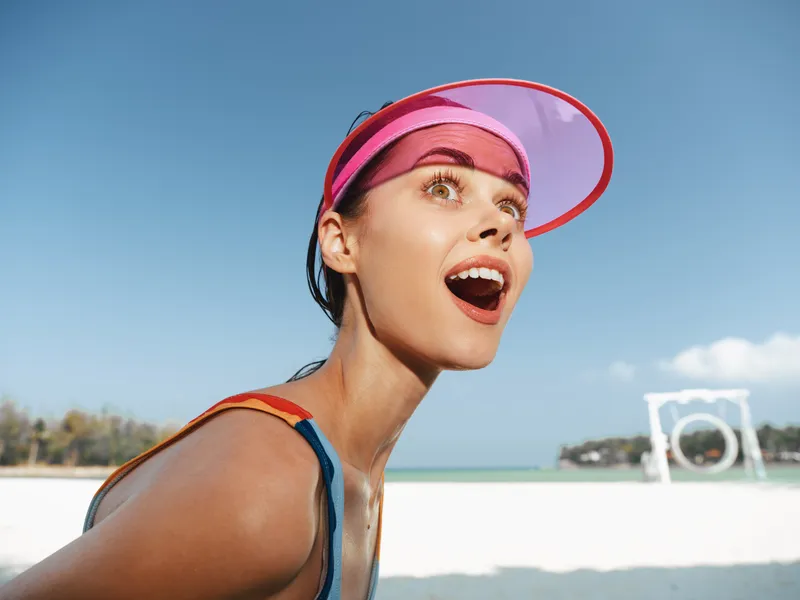 Dynamic close up of a joyful woman on a sunny beach, wearing a pink visor, wide smile, and bright eyes, capturing energy and carefree summer mood.