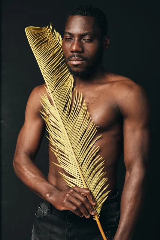 African man with dark skin holding large golden palm leaf in creative conceptual visual style. Shirtless male model posing with closed eyes in artistic studio portrait with black background.