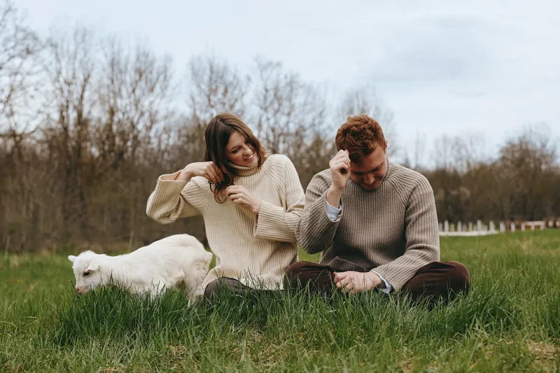 A couple sits on bright green grass in an open meadow, a curious goat kid near them. They smile and interact with the animal, capturing a warm, relaxed rural lifestyle moment.