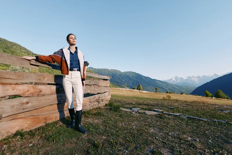 Woman in jacket and boots standing by a wooden fence in countryside meadow with mountains in background, embracing nature and wide landscape in a sunlit outdoor relaxed scene for lifestyle.