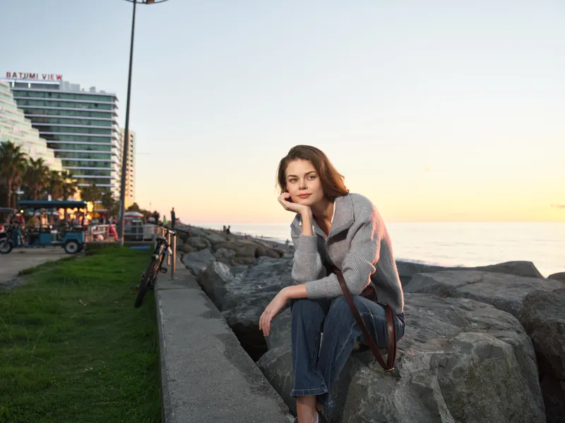 A thoughtful woman sits on coastal rocks at sunset, gazing toward the horizon. She wears casual attire and a crossbody bag, enjoying a peaceful moment by the sea, with warm light bathing the scene.