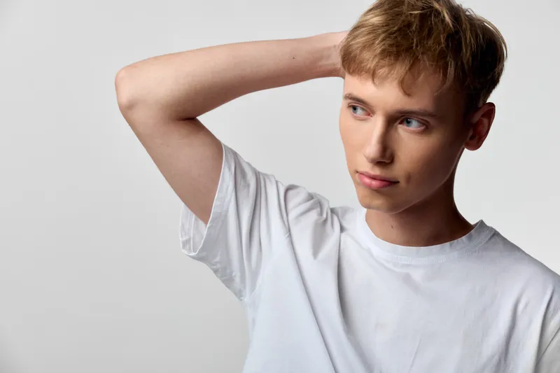 Man in a casual white shirt stands in a light studio, touching the back of his head with a thoughtful expression, conveying introspection, curiosity, and subtle emotion for lifestyle or fashion