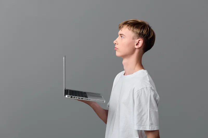 Youthful student holding a laptop in one hand, showcasing technology, focus and learning in a minimal studio with neutral grey background and clean white tshirt