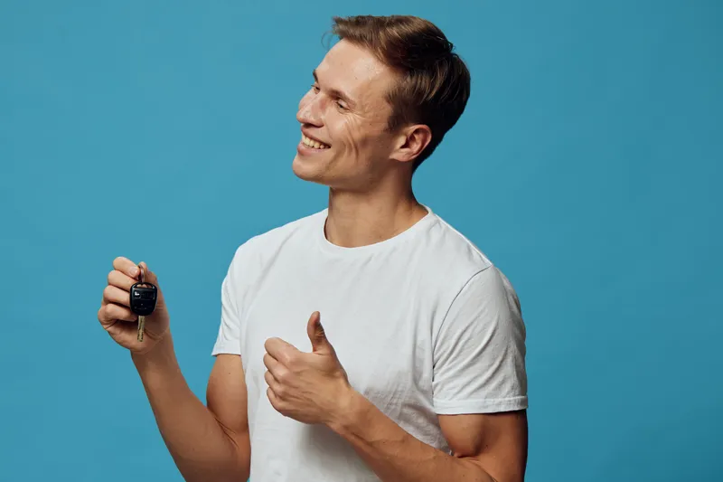 Smiling man with short brown hair wearing casual white t-shirt holding a remote control and giving a thumbs up on bright blue background, studio portrait. People lifestyle concept