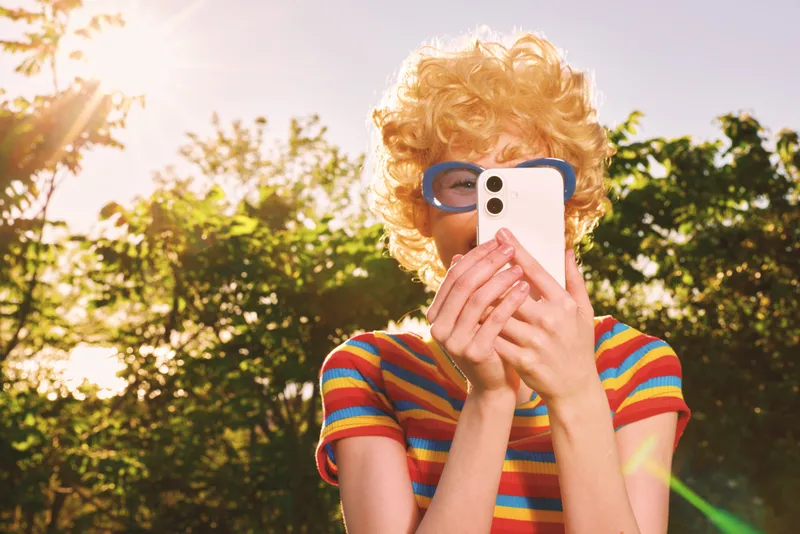 Young woman with a blonde curly wig takes a playful selfie in a sunlit park, wearing a striped shirt and oversized sunglasses, channeling retro film vibes and carefree mood.