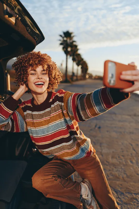Smiling woman with curly hair taking a selfie with smartphone beside a car, outdoor road trip, sunset sky and palm trees, casual striped sweater, lifestyle happiness