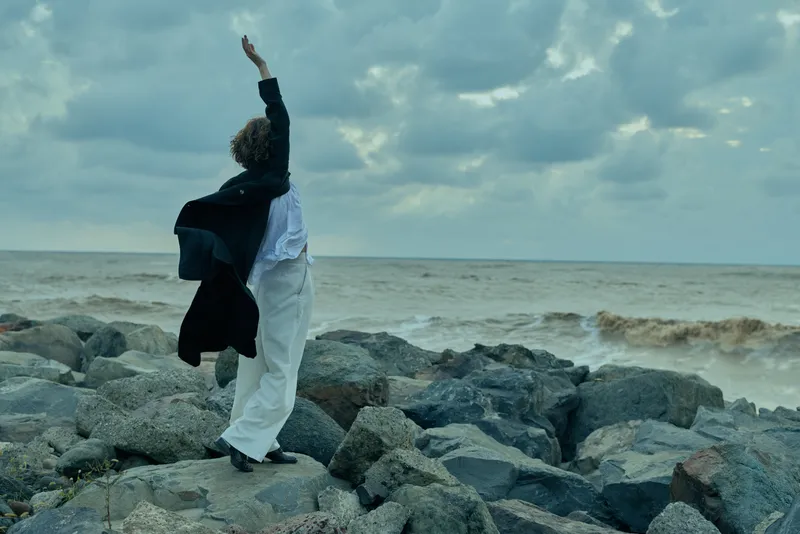 Figure on rocky shoreline with waves, wind blowing coat, raised arm toward cloudy sky, expansive sea horizon, dramatic outdoor mood and motion in a blue gray seascape