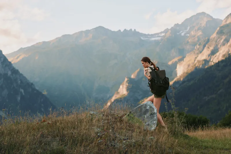A young woman with a backpack hikes along a grassy alpine trail, framed by distant mountains under a clear blue sky, appearing confident and focused with a sense of adventure and freedom.