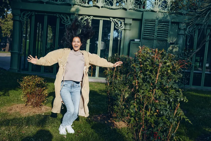 Girl jump outdoor joy smile yard garden coat jeans laughter and a carefree moment captured during daylight, conveying energetic motion and playful happiness in a candid scene