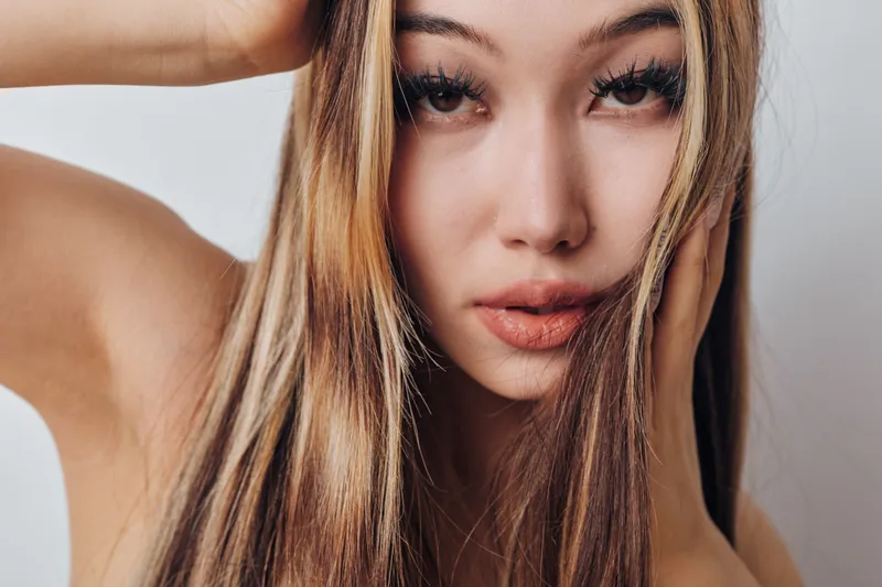Young woman with long blonde hair posing closeup with natural makeup and serious expression on light background. Beauty, portrait, female model with clear skin and hand near face.