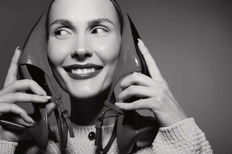 Smiling white woman with light skin holds two high-heeled shoes near her face. Black and white studio portrait with playful and confident expression, fashion concept.