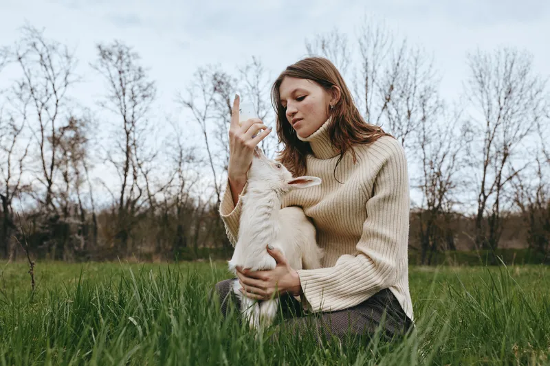 A woman in a beige sweater sits in a grassy field, cradling a young goat kid in her arms, with a focused gaze. A lifestyle scene highlighting animal companionship, care, and bonding outdoors.