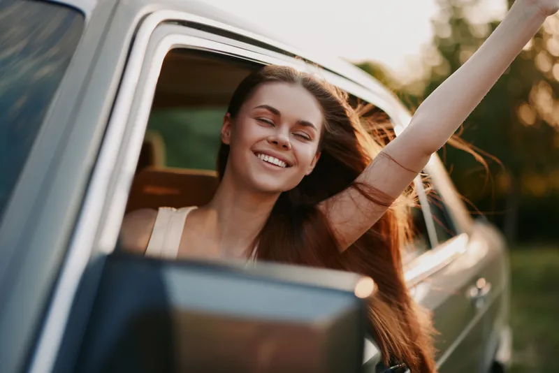 Happy young woman with long hair enjoying a road trip, smiling while leaning out of a car window during sunset, conveying freedom and joy.