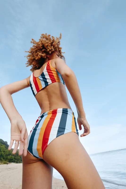 Young woman with curly hair wearing colorful striped swimsuit standing on beach under blue sky. Summer vacation, ocean, and outdoor lifestyle concept with natural light and relaxed mood.
