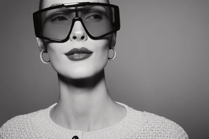 Close-up portrait of a fair skinned Caucasian woman wearing oversized sunglasses and hoop earrings with a knitted sweater on light background.