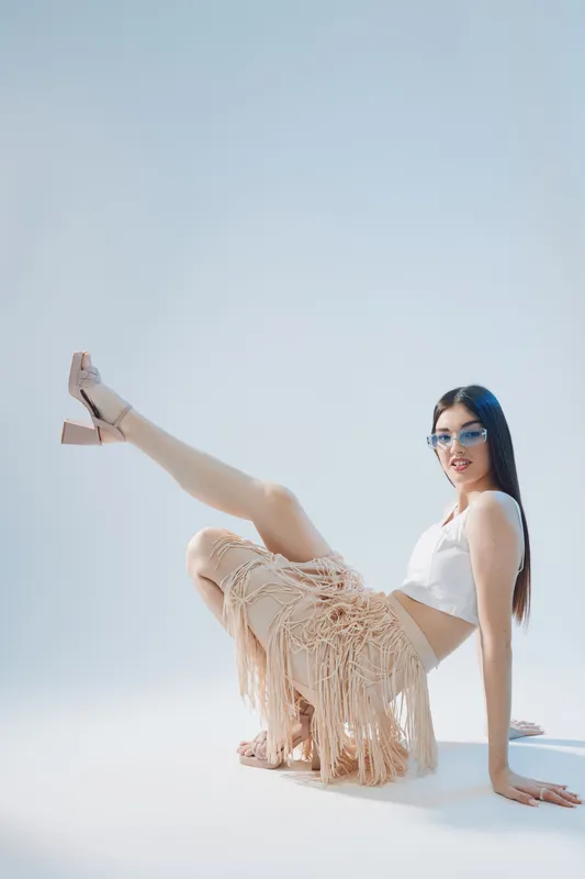 Young woman in a white top and beige fringed skirt poses with one leg raised on a light background. Concept of beauty, fashion, and confident style.