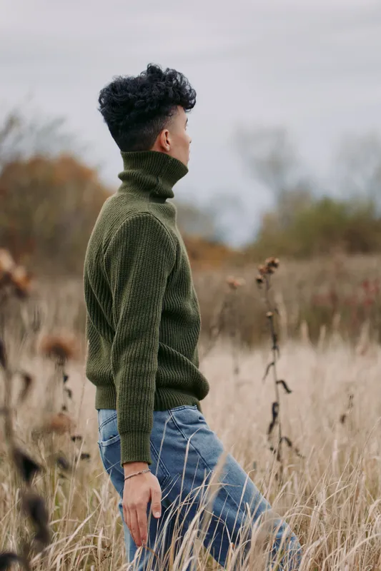 Young man in an olive knit sweater stands in a dried meadow, captured in an authentic, realistic candid portrait conveying quiet reflection, natural posture, textured clothing and soft overcast light.