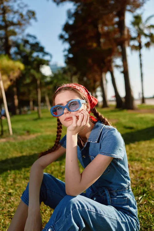 Young woman with braids wearing blue sunglasses and denim outfit sits on grass in sunny park during afternoon, relaxing with thoughtful expression and casual style.