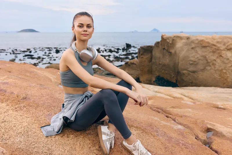 Young fit woman in activewear relaxing on rocky beach, headphones on, showcasing a healthy lifestyle and serene emotions amidst a coastal background