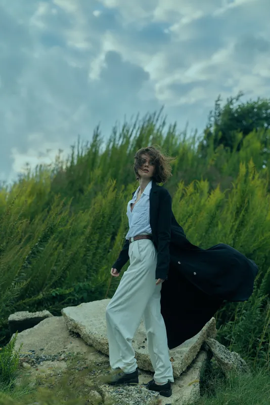 Fashionable woman in a long black coat over white suit steps on rocky terrain with lush greenery, capturing bold movement and cinematic style in an outdoor wind swept environment