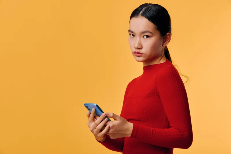 Young woman in red sweater holding smartphone and looking at camera with serious expression on yellow background. Modern technology, communication and casual style portrait with copy space.