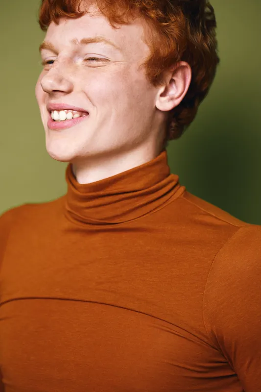Young man with red hair wearing orange turtleneck smiling with eyes closed in studio. Casual portrait of happy male model, healthy skin and natural expression on green background.