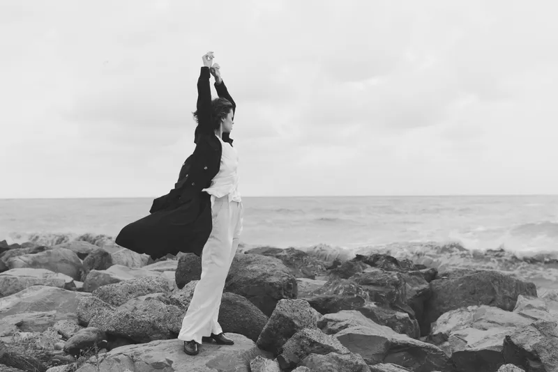 Woman stretches arms overhead on a rocky shore, dress flowing, by the sea, conveying freedom, wind, and outdoor resilience in a monochrome seascape.