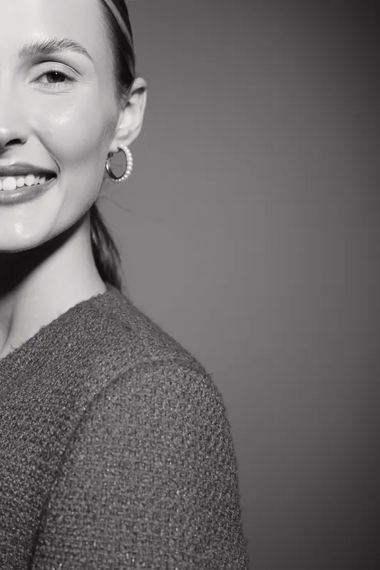 Smiling Caucasian woman with light skin and hoop earrings wearing textured sweater. Close-up portrait in black and white with soft lighting on plain background. Concept of happiness and style.