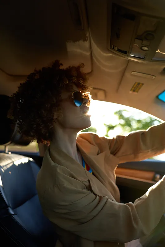 A woman drives a car in a casual lifestyle moment, wearing sunglasses as sunlight glows through the windshield, with a retro film color vibe and warm urban mood.