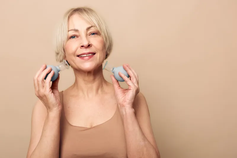 Smiling middle aged woman with short blonde hair holds two facial cleansing brushes near face on beige background. Concept for skincare, beauty routine, self care, and healthy skin.