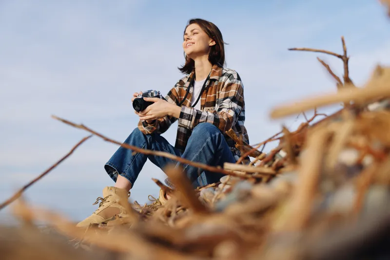 Photography of a woman photographer outdoors, sitting among branches with a camera, wearing a plaid shirt and jeans, enjoying nature and a peaceful landscape moment.