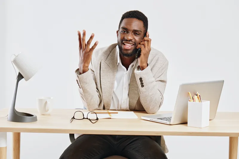 Confident smiling man talking on phone at desk with laptop and notebook in modern home office setting, casual clothing, positive mood, work from home environment, indoor lifestyle, cheerful profession