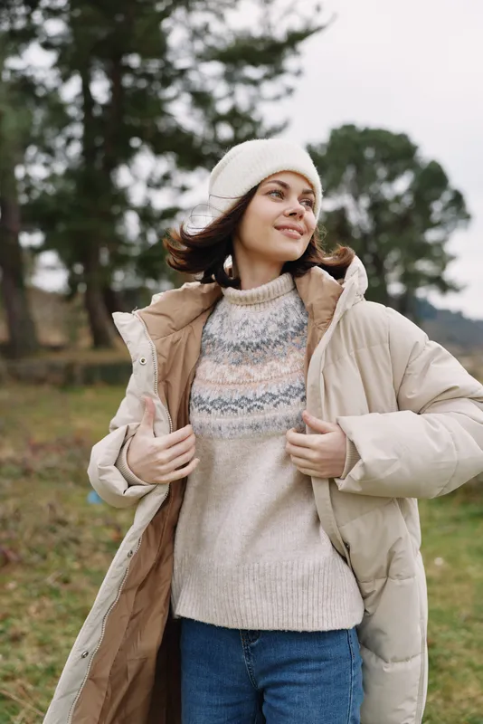 Woman in cozy winter outfit posing outdoors in a park, wearing a knit sweater, puffy coat and beanie, confident expression, soft natural light and autumnal scenery.