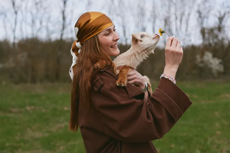 A woman in a brown coat and headscarf gently holds a playful goat kid, smiling in an open field, capturing a lifestyle moment of care, bonding, and rural life with animals.