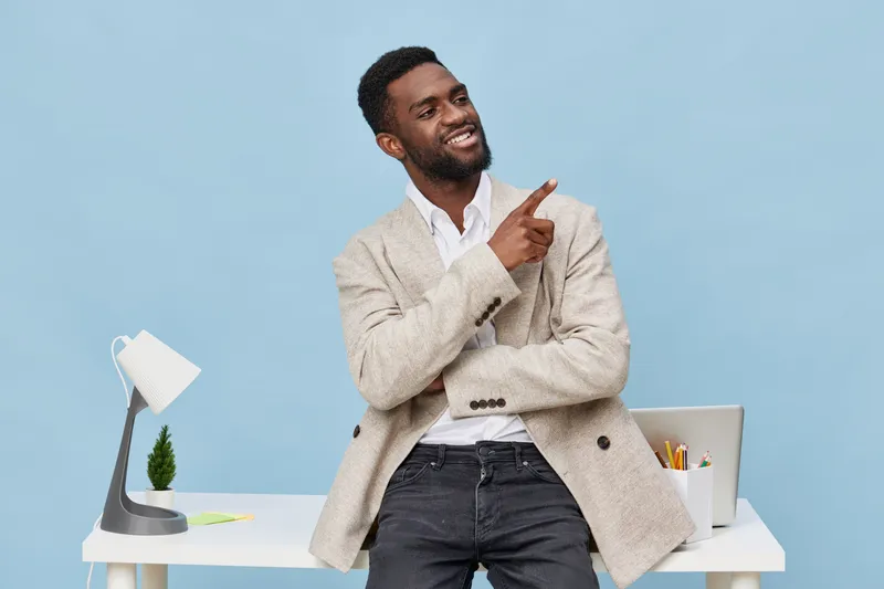 Confident smiling man with dark skin, wearing a beige blazer and white shirt, standing in modern office setting with blue background, pointing upwards, professional and positive mood. People lifestyle