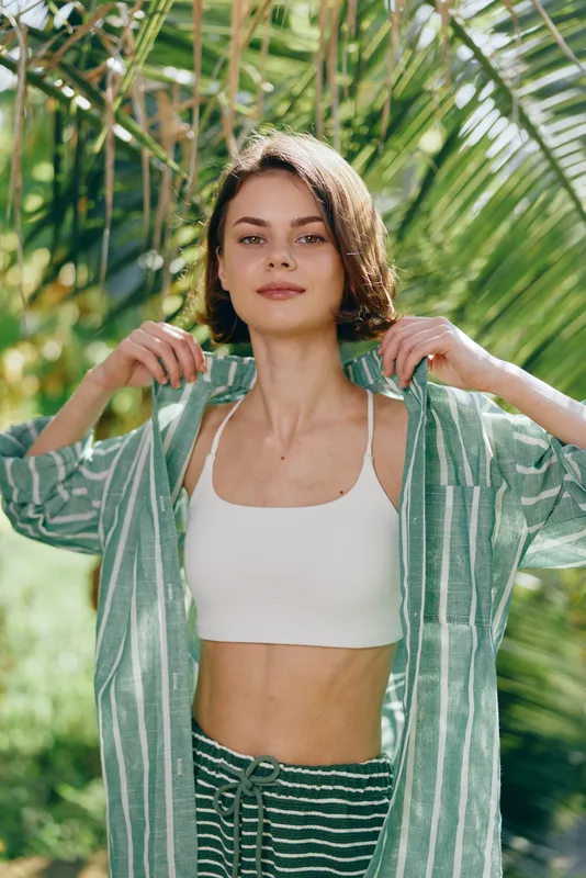 Portrait of a young woman wearing a white crop top under a green striped coverup, posing outdoors amid tropical foliage.
