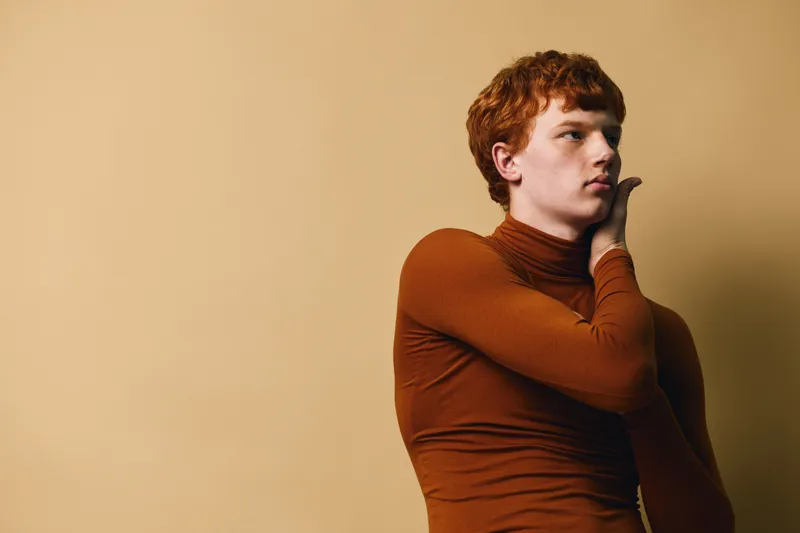 Young man with red hair wearing brown turtleneck touching face with hand looking sideways. Studio portrait with beige background showing thoughtful expression and casual style fashion.